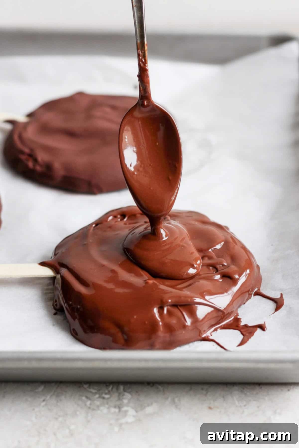 Delicious Homemade Dilly Bars 4 A spoon drizzles melted chocolate onto a round treat on a parchment-lined tray with a popsicle stick.