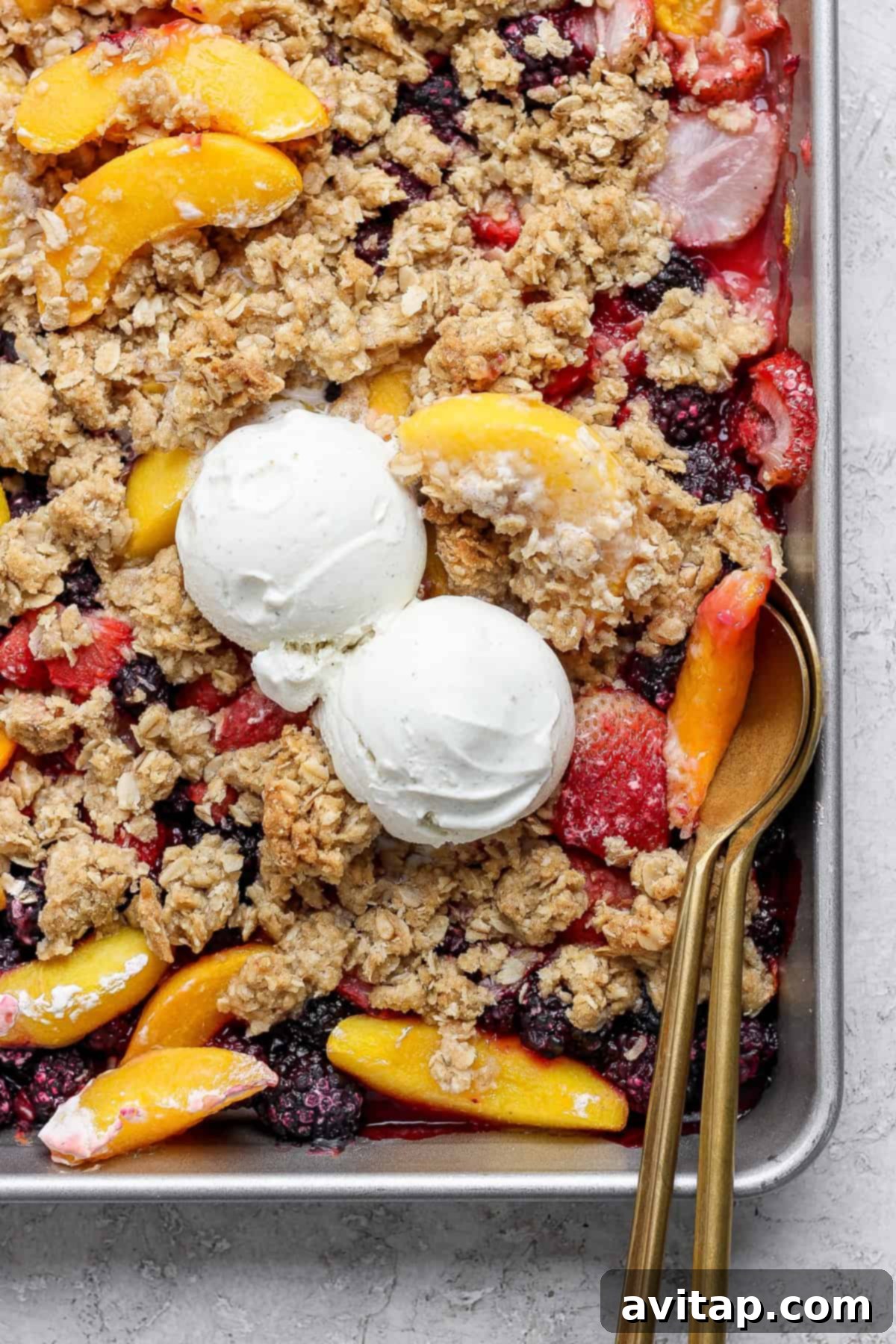 A tray of mixed fruit crumble topped with two scoops of vanilla ice cream and oat crumbles, with a serving spoon on the right side.