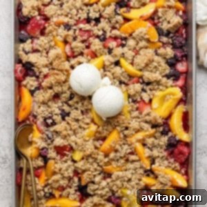 A rectangular baking tray filled with a mixed berry and peach crumble topped with two scoops of vanilla ice cream. A gold spoon rests on the left side of the tray.