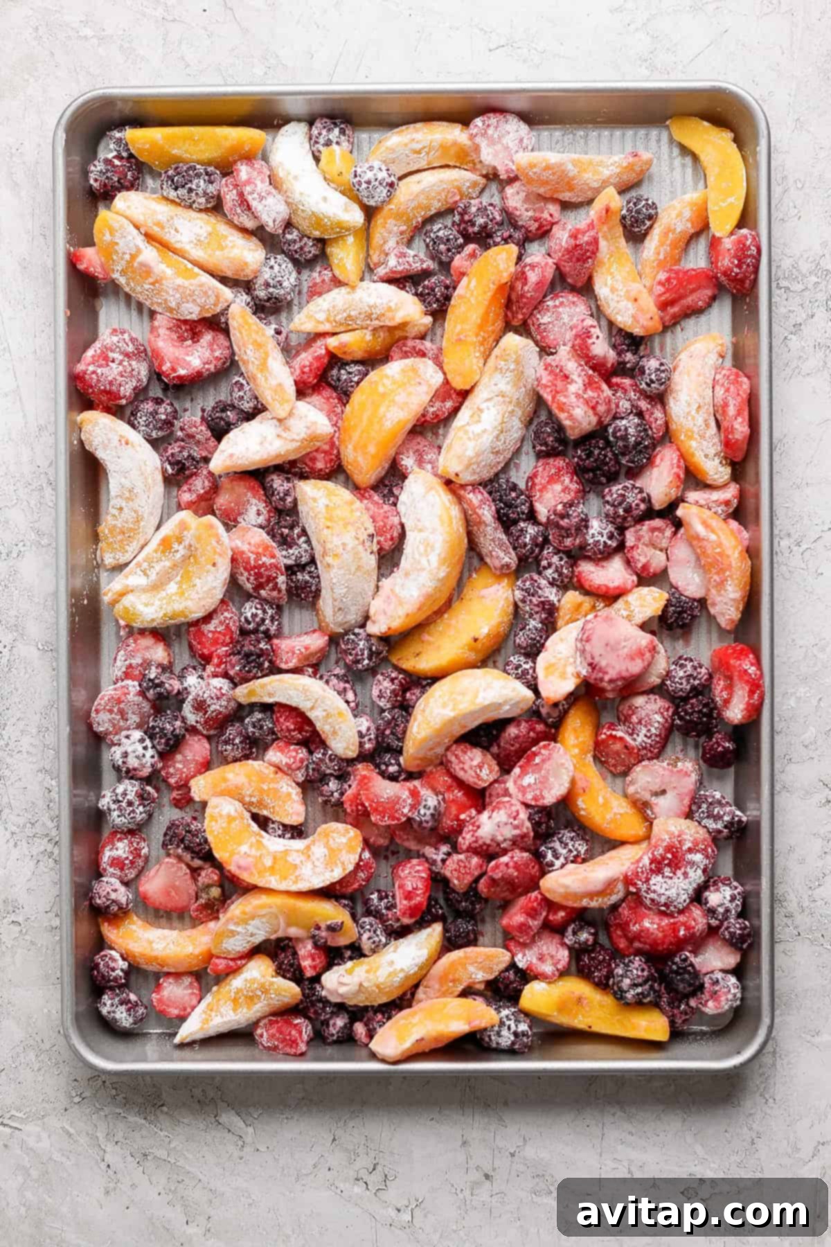 A baking sheet filled with an assortment of frozen fruit slices including peaches, strawberries, and blackberries arranged evenly across the surface.