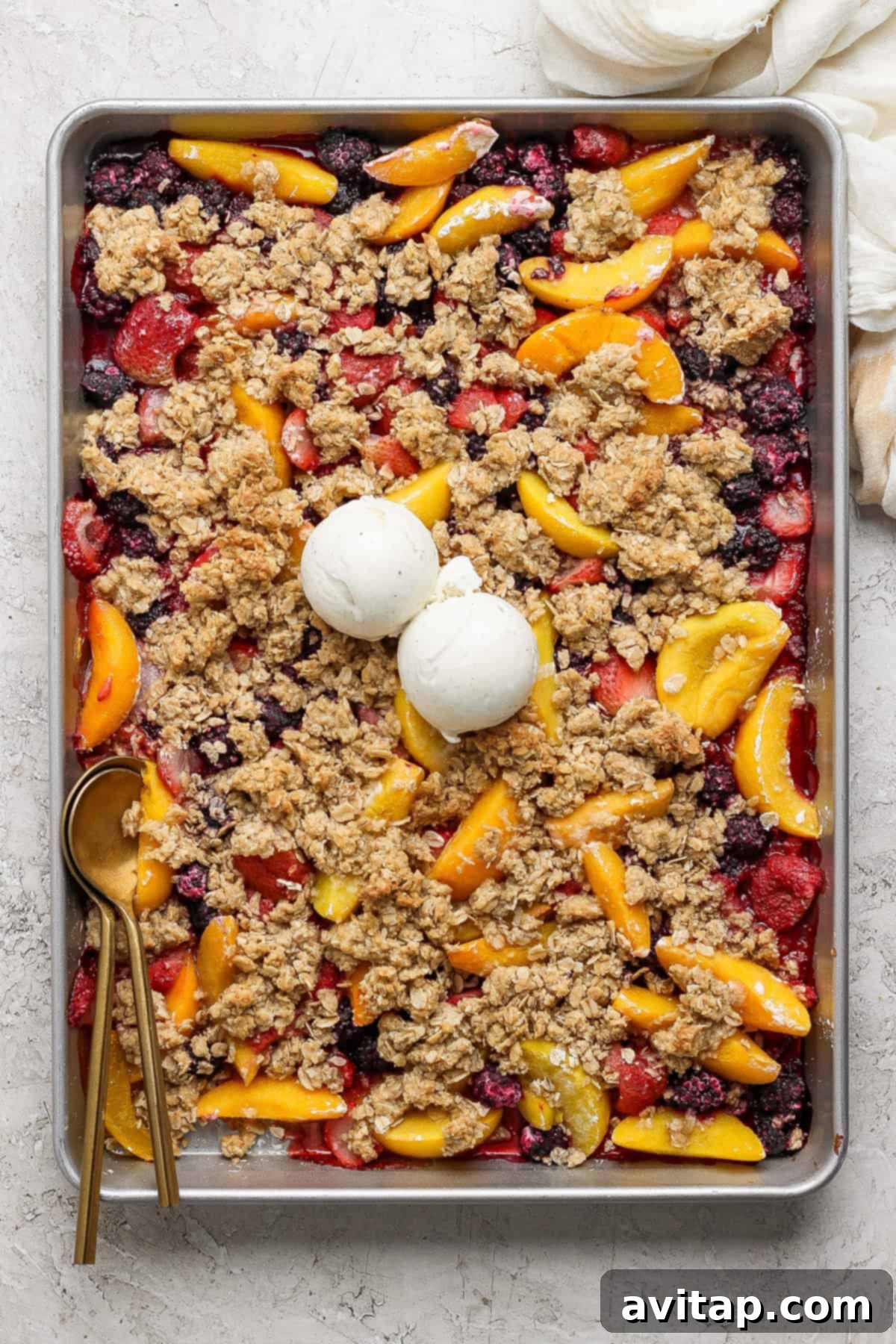 A rectangular baking tray filled with a mixed berry and peach crumble topped with two scoops of vanilla ice cream. A gold spoon rests on the left side of the tray.