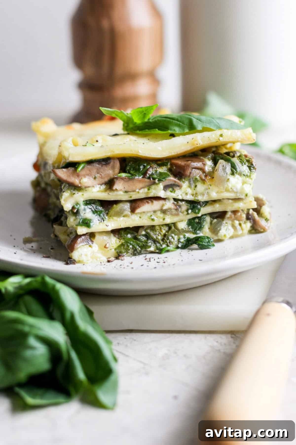 Artisan Pesto Lasagna 2 A perfectly portioned slice of vegetarian pesto lasagna on a white plate, showcasing its distinct layers of pasta, vibrant green pesto, creamy cheese, sautéed spinach, and mushrooms, garnished with fresh basil.