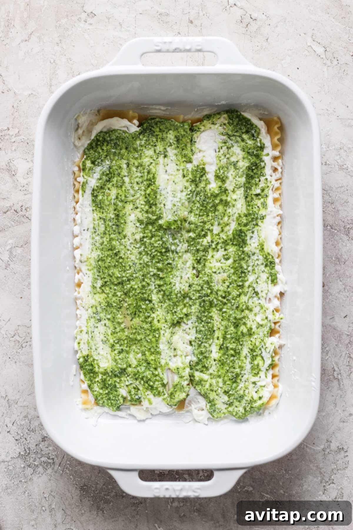 Artisan Pesto Lasagna 4 An uncooked pesto lasagna layered in a white rectangular baking dish, showing visible green pesto, white cheese mixture, and golden pasta layers, set against a light gray textured background.