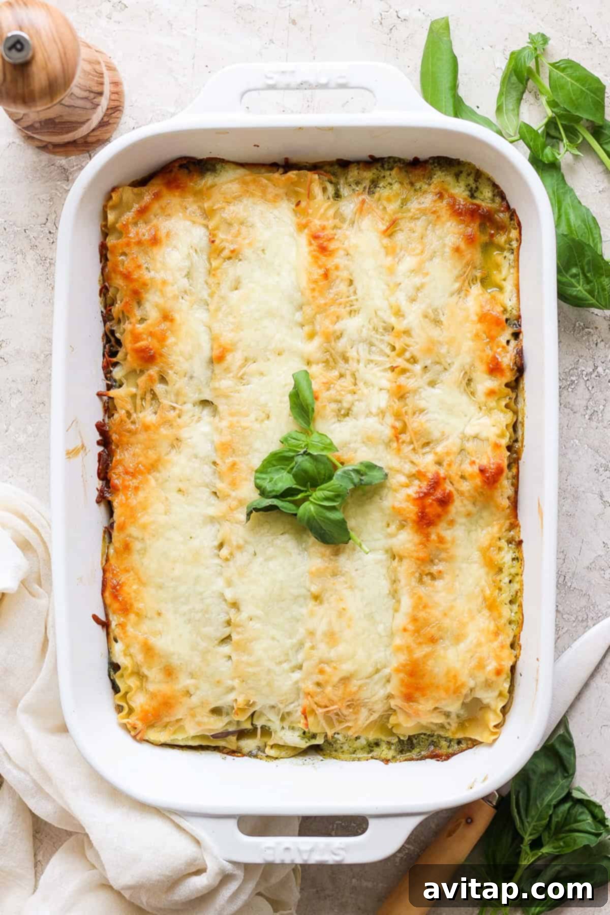 Artisan Pesto Lasagna 5 A freshly baked pesto lasagna in a white rectangular baking dish, topped with golden melted cheese and vibrant fresh basil leaves. A wooden pepper grinder and additional basil leaves are visible in the background.