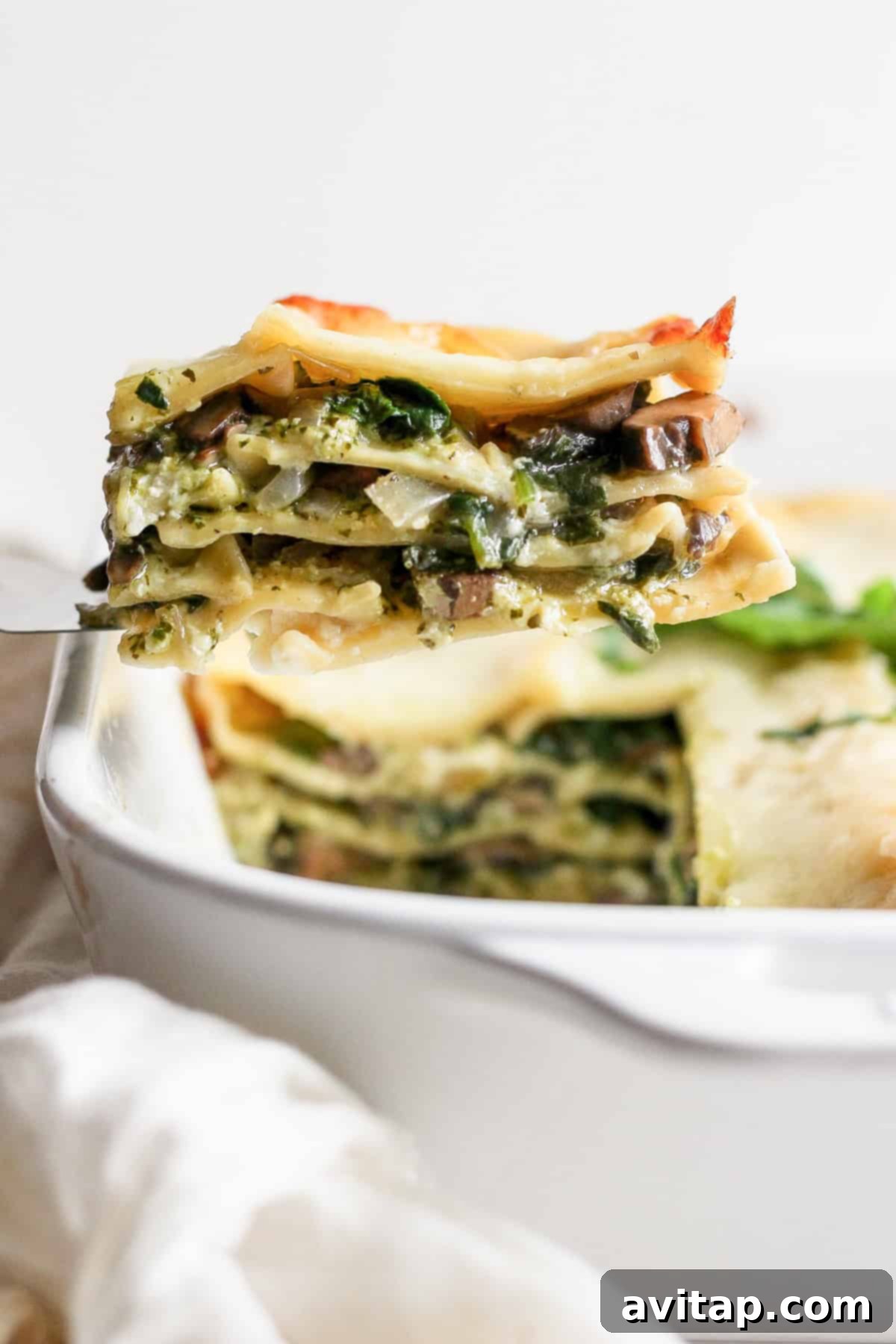 Artisan Pesto Lasagna 7 A tantalizing close-up of a slice of vegetable pesto lasagna being lifted from a white baking dish, revealing perfect layers of pasta, green pesto, creamy cheese, and sautéed vegetables.