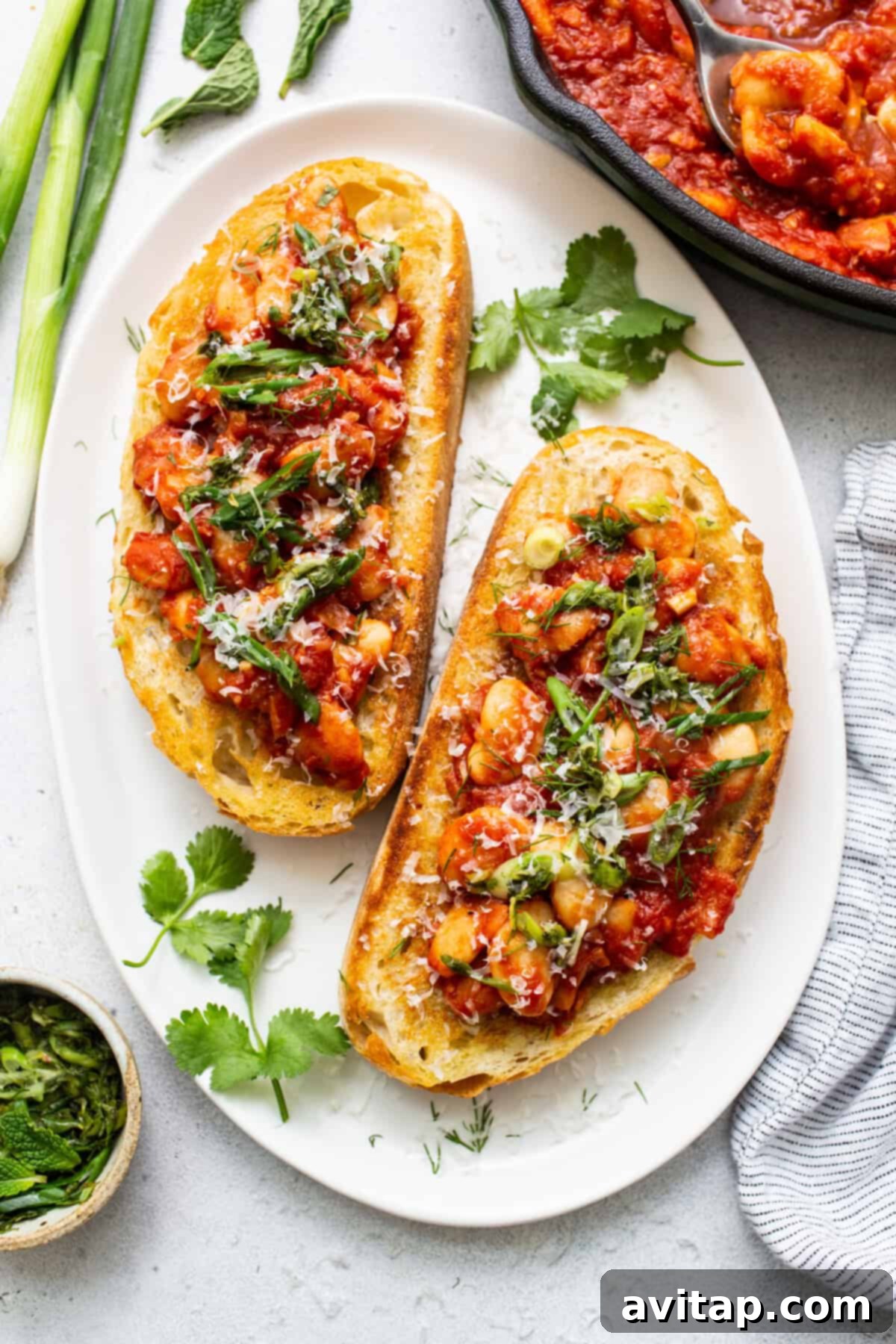 Two slices of hearty toast generously topped with a rich, vibrant tomato and butter bean mixture, garnished with fresh, green herbs and a sprinkle of cheese.