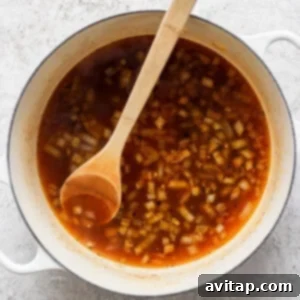A white pot filled with a red broth containing chopped onions, stirred with a wooden spoon, placed on a gray textured surface.