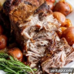 A close-up of a seasoned, cooked roast surrounded by whole red potatoes and fresh rosemary sprigs on a white platter. A fork and knife are set to the side.