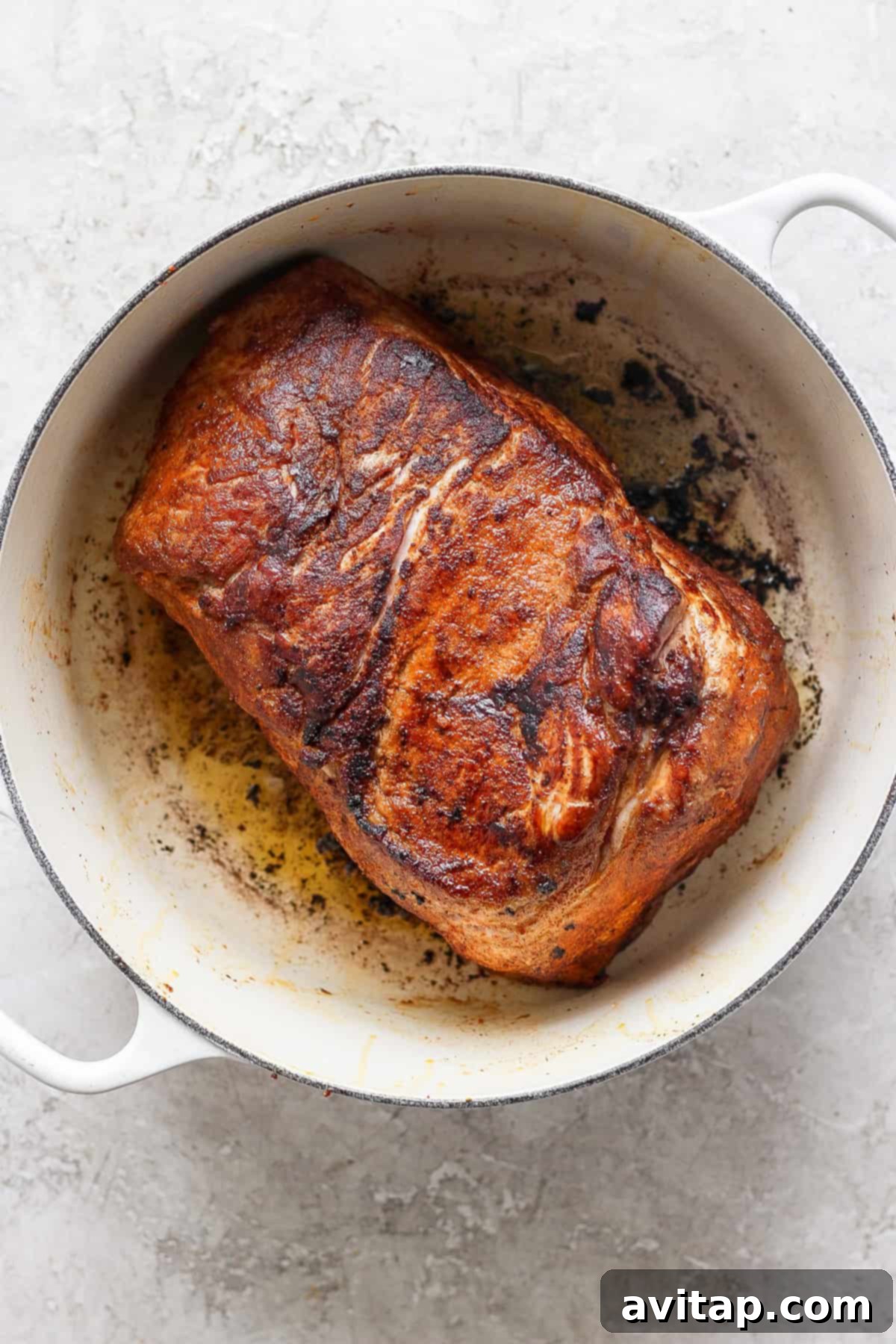 A seared, beautifully browned piece of pork shoulder rests in a white enameled cast iron Dutch oven, displaying its crispy, caramelized edges.