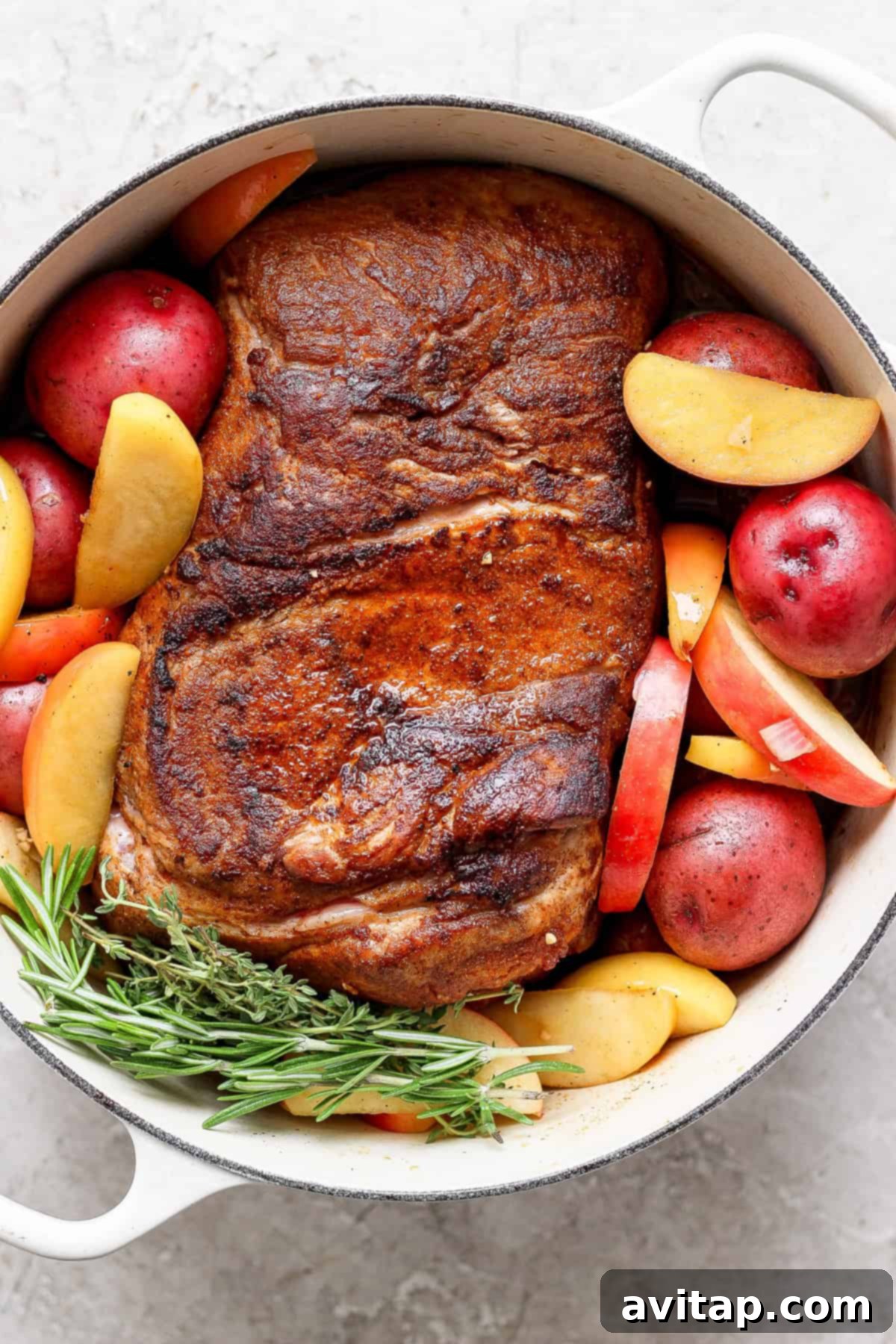 A cooked pork roast with a perfectly crispy exterior, surrounded by tender red potatoes, thinly sliced apple pieces, and fragrant sprigs of rosemary, all nestled within a pristine white cast iron Dutch oven.