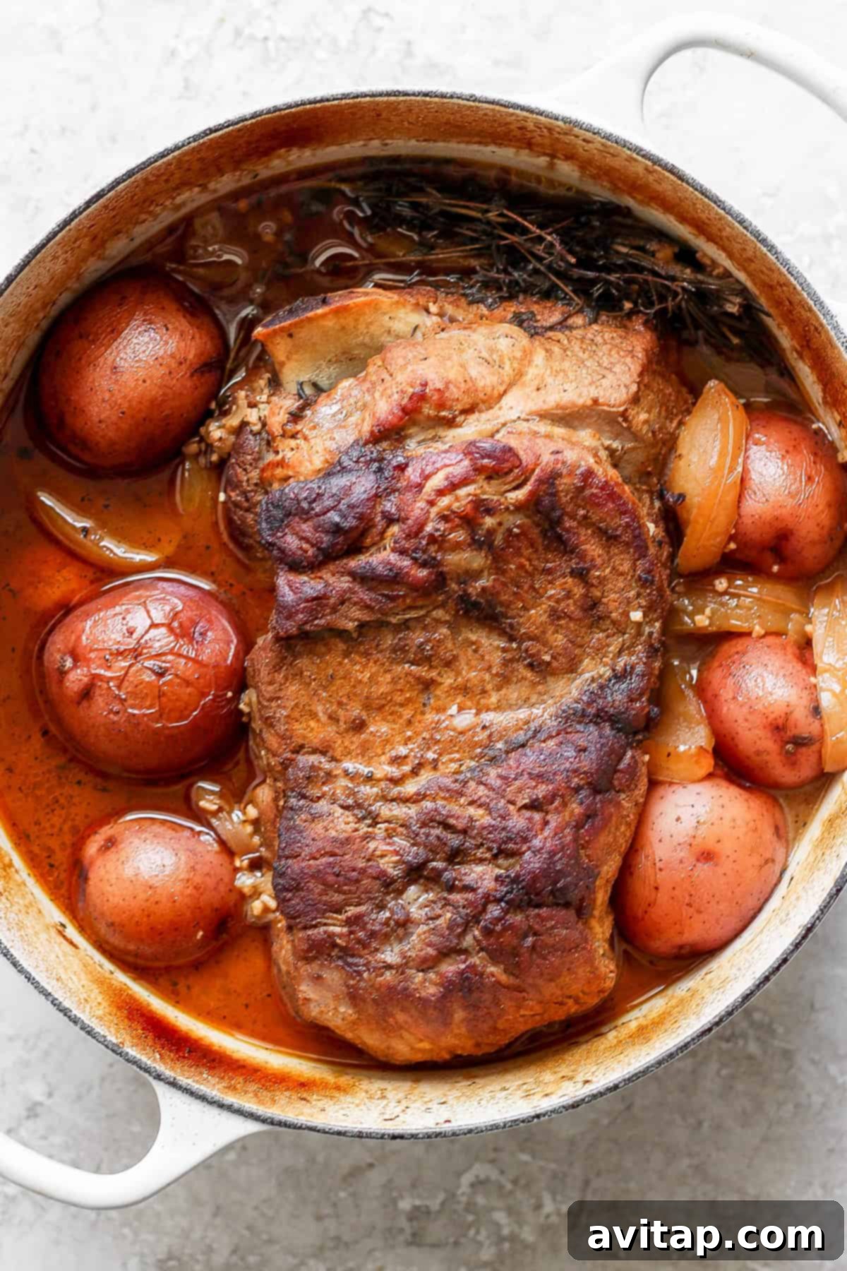 A cooked pot roast, surrounded by tender potatoes and onions, resting in a pristine white enameled cast iron pot, showcasing the rich, savory sauce.