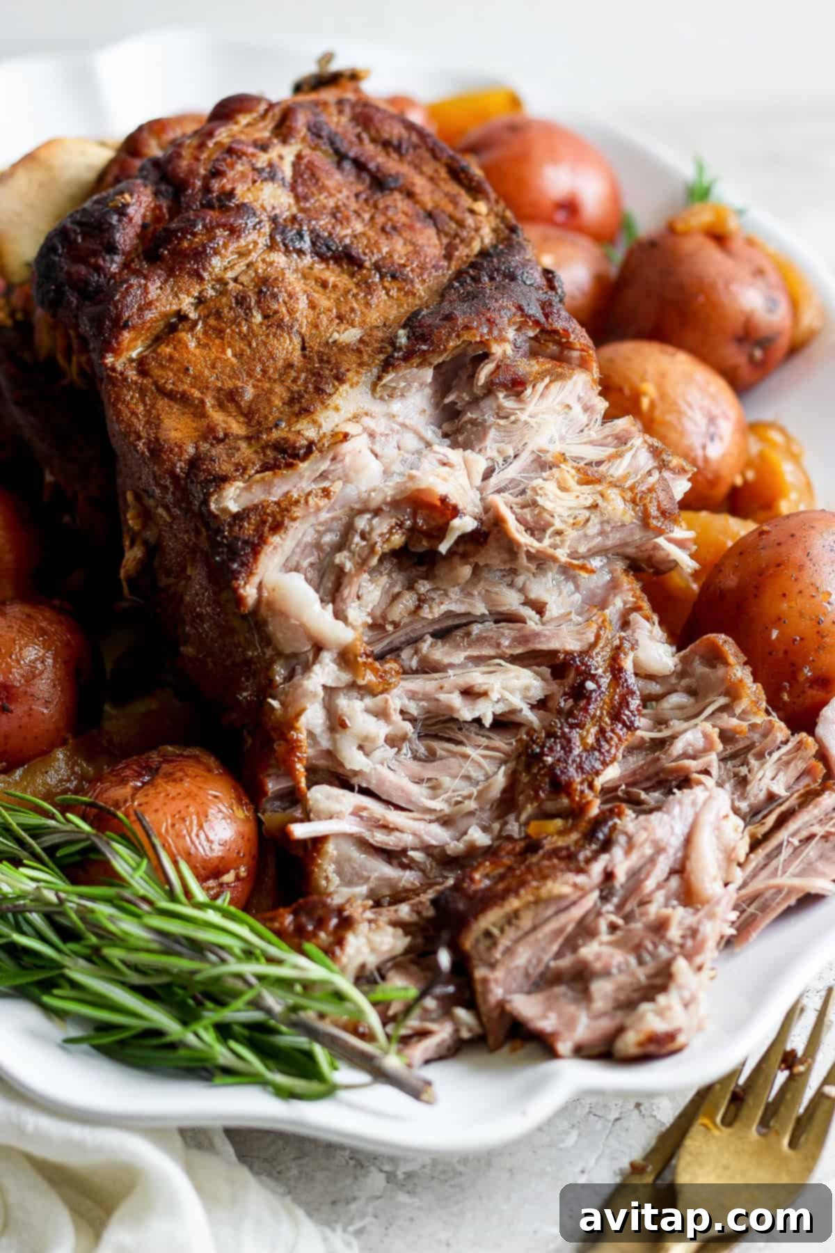 A close-up view of a perfectly seasoned and cooked pork roast, beautifully surrounded by whole baby red potatoes and fragrant fresh rosemary sprigs, presented on a pristine white platter. A fork and knife are artfully arranged to the side, signaling readiness for serving.