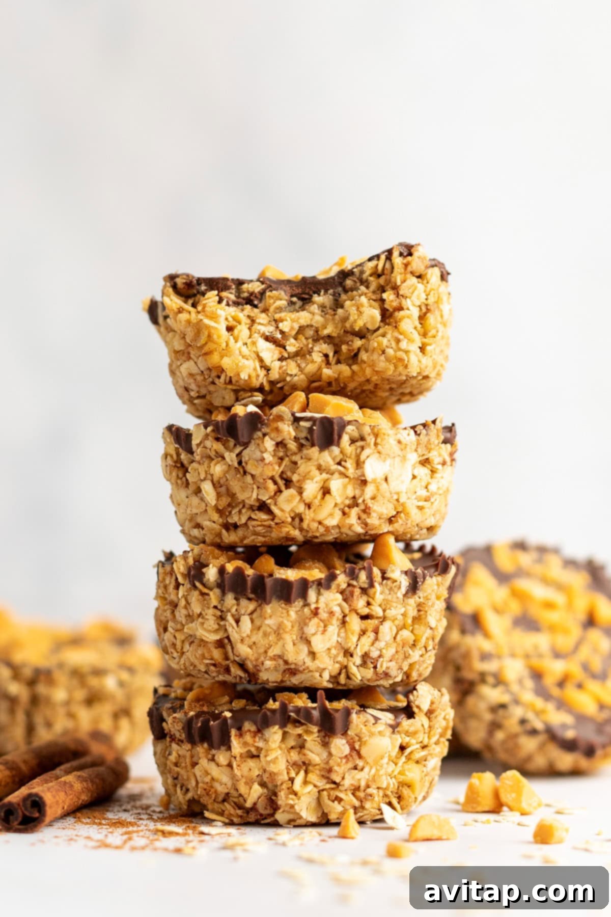 A beautifully arranged stack of four no-bake pumpkin spice oat cups, featuring a rich chocolate topping and a visible bite taken from the top cup, showcasing the textured oat base. The cups are presented on a light surface, surrounded by scattered autumn-themed ingredients like walnuts and pumpkin spice, evoking a cozy fall atmosphere.