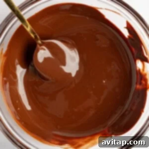 A simple white bowl filled with glossy, freshly melted chocolate, with a spoon resting inside it. The smooth texture of the chocolate suggests it has been perfectly melted and is ready to be used as a topping or dip.