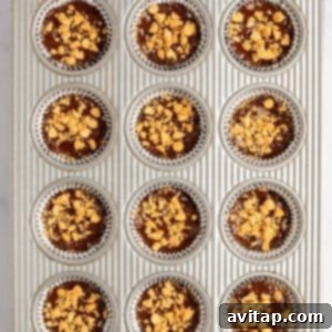 A shiny metal muffin tin, showcasing twelve individual slots filled with a dark, rich chocolate batter. Each slot is lined with a silver foil cupcake liner, and the chocolate topping is generously adorned with crushed nuts, making the cups ready for chilling or the next stage of preparation.
