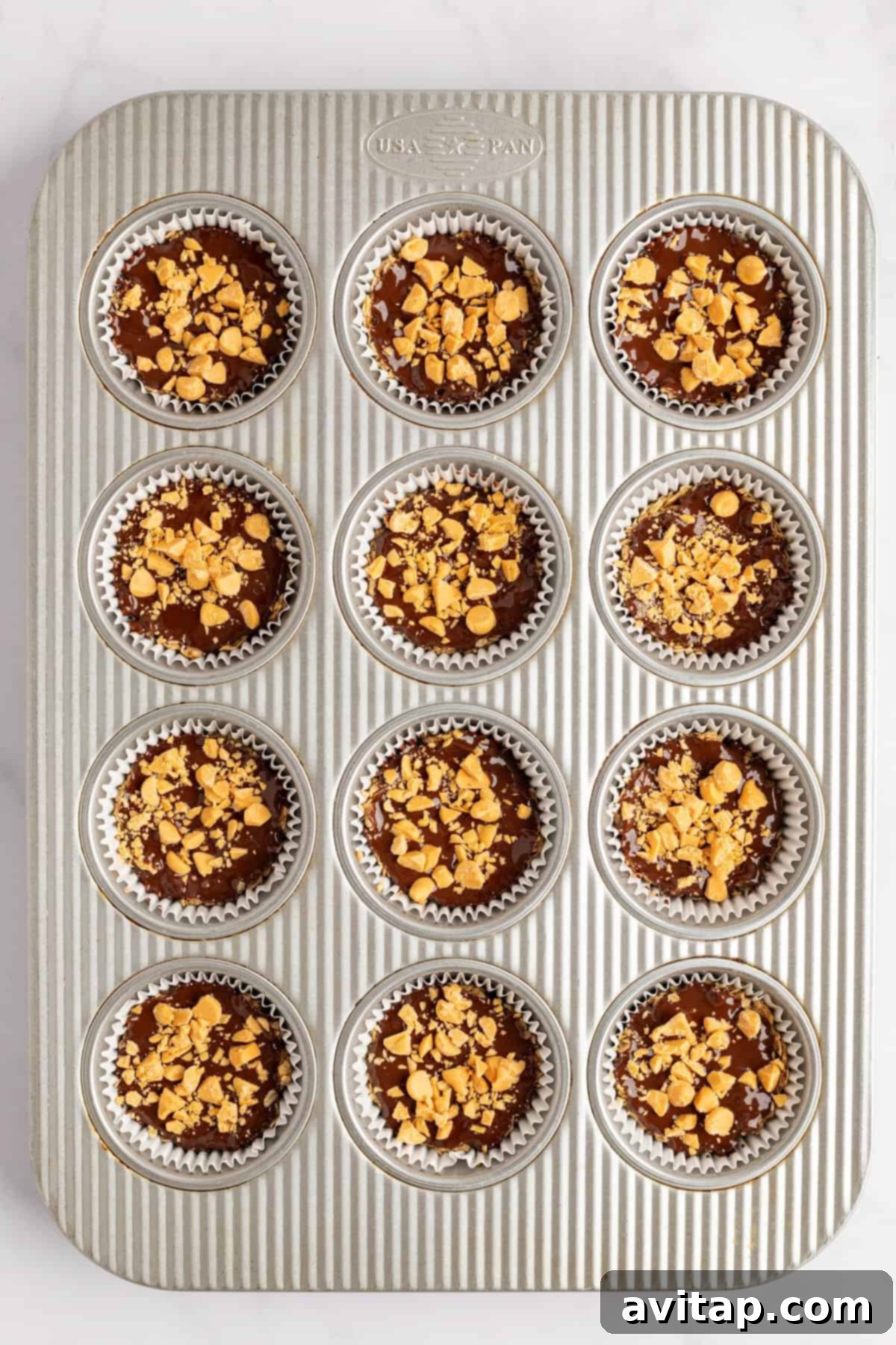A standard metal muffin tin with twelve slots, each lined with a silver foil cupcake liner. The liners are filled with a dark, rich chocolate batter and topped generously with crushed nuts, suggesting they are ready for the next stage of preparation or chilling.
