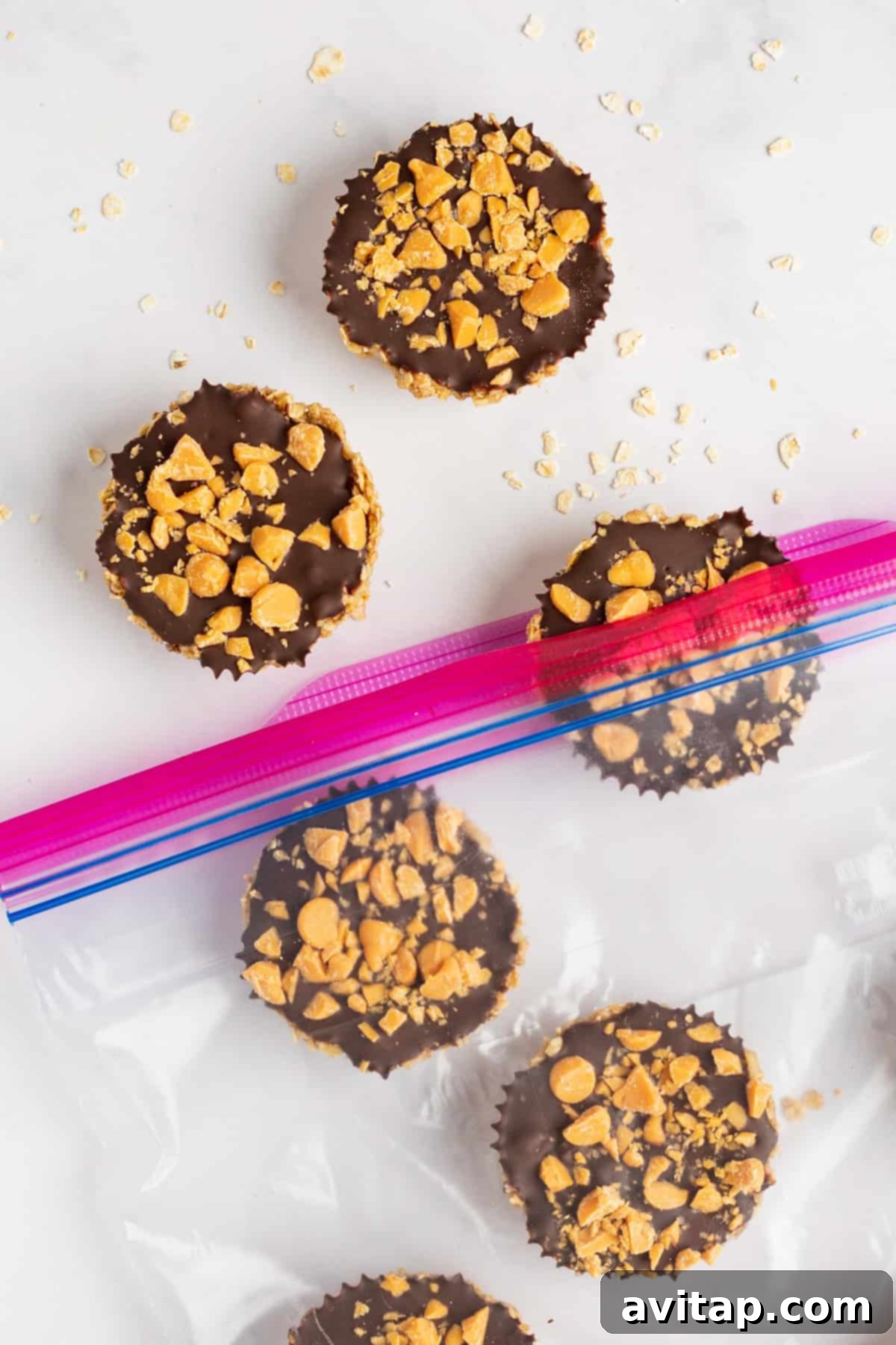 A close-up shot of five homemade no-bake treats, resembling chocolate peanut butter cups. Each cup features a rich chocolate topping generously sprinkled with crushed peanuts. Three cups are neatly arranged inside a partially open resealable bag, while two rest outside on a clean white surface, surrounded by scattered peanut pieces.