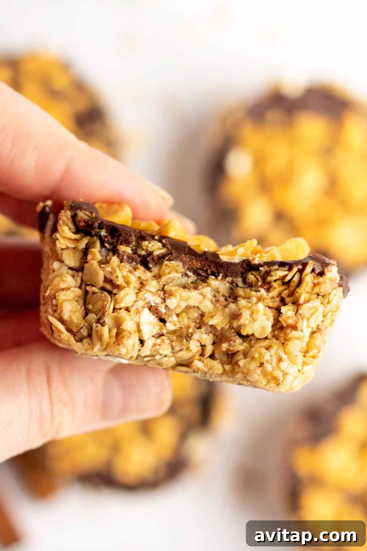 A close-up shot of a hand delicately holding a homemade granola bar with a rich chocolate topping, artfully sprinkled with various nuts. In the background, several more granola bars are visible, slightly out of focus, creating a sense of abundance and deliciousness.