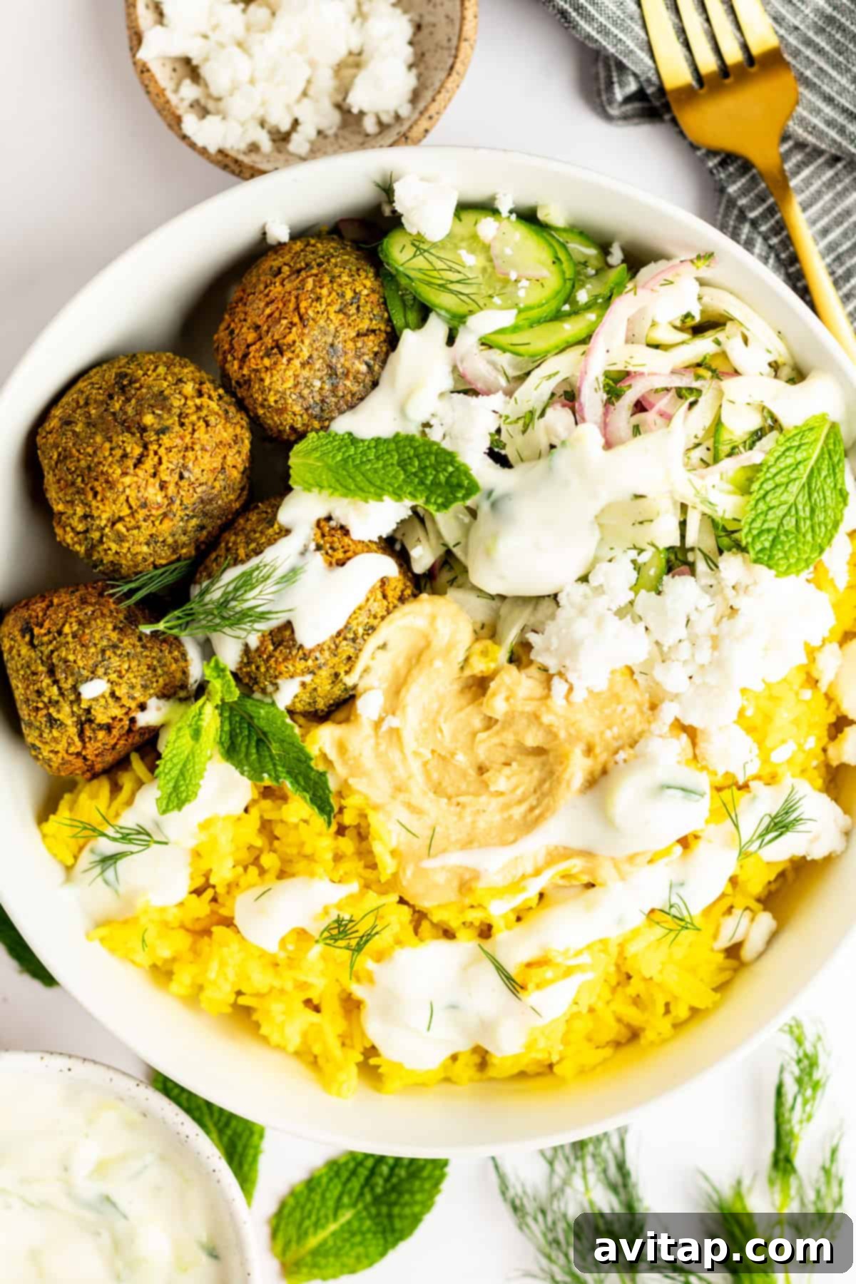 A beautifully arranged falafel bowl featuring golden falafel, fluffy yellow turmeric rice, fresh cucumber salad, creamy hummus, crumbled feta cheese, and fragrant herbs, all drizzled with a white sauce. A gold fork rests beside the bowl on a grey napkin.