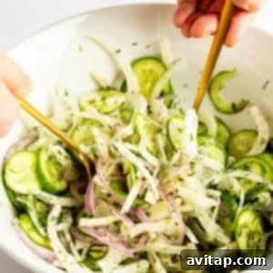 Hands mixing a salad with cucumbers, red onions, and herbs in a white bowl using two gold utensils.