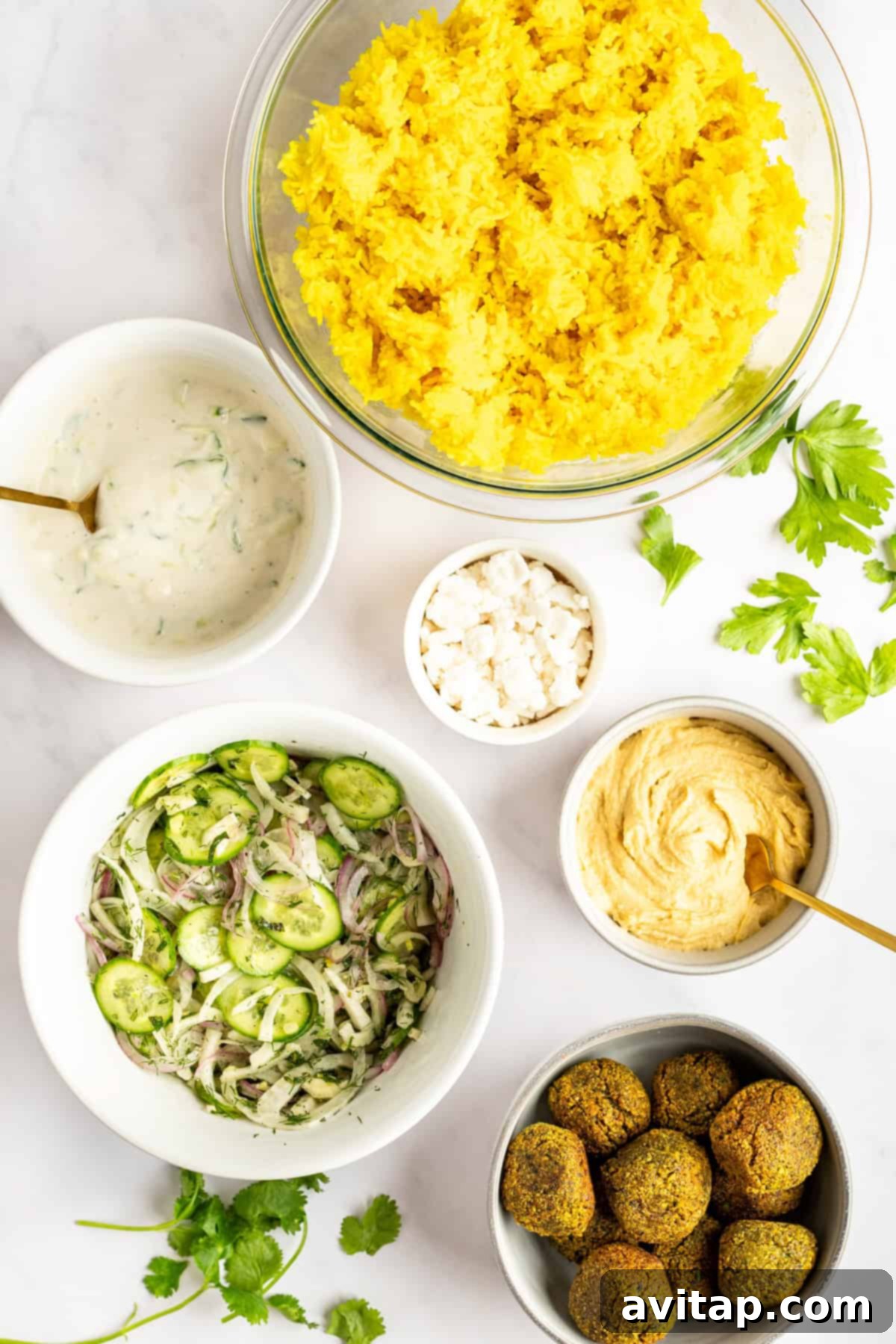 An array of freshly prepared falafel bowl components laid out on a white surface, including a bowl of yellow turmeric rice, a small dish of creamy yogurt sauce, crumbled feta cheese, a serving of hummus, a vibrant cucumber salad, and golden-brown falafel patties, all garnished with sprigs of cilantro.