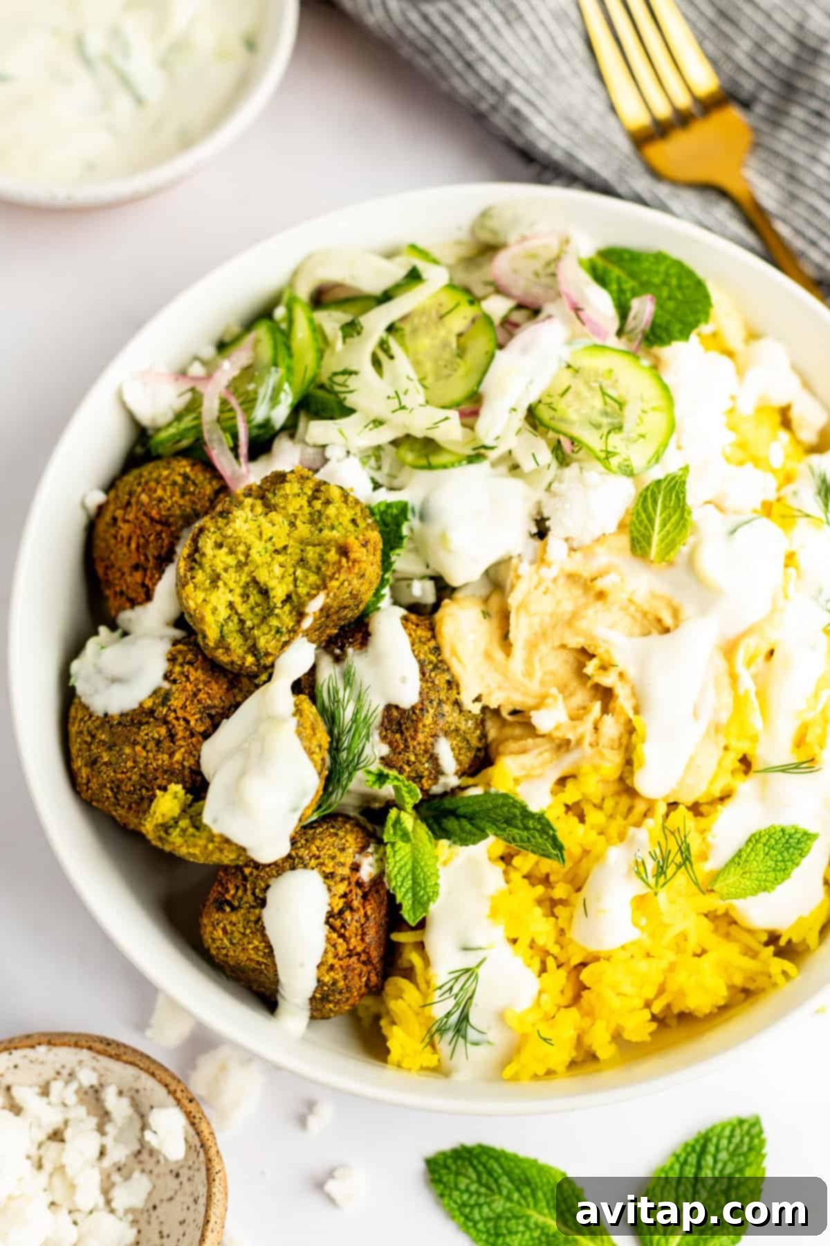 A close-up shot of a prepared falafel bowl, featuring yellow turmeric rice, golden falafel patties, thinly sliced cucumbers and red onions, fresh dill, crumbled feta cheese, and mint leaves, all topped with a dollop of hummus and a drizzle of white sauce. A fork and a small bowl of dip are visible in the soft background.