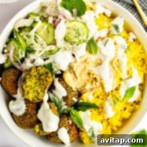A bowl containing yellow rice, falafel, sliced cucumber, red onions, dill, feta cheese, and mint leaves, topped with hummus and drizzled with a white sauce.