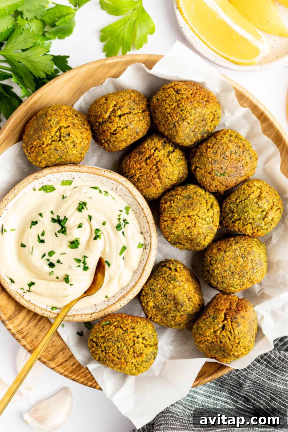 A wooden plate holds ten falafel balls surrounding a bowl of creamy dip garnished with herbs. Lemon wedges and parsley leaves are in the background.