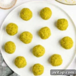 A white plate with twelve round, greenish-yellow falafel balls. Nearby are a bowl of falafel mixture, a bowl of salt, three garlic cloves, a cloth, and a pair of tongs.