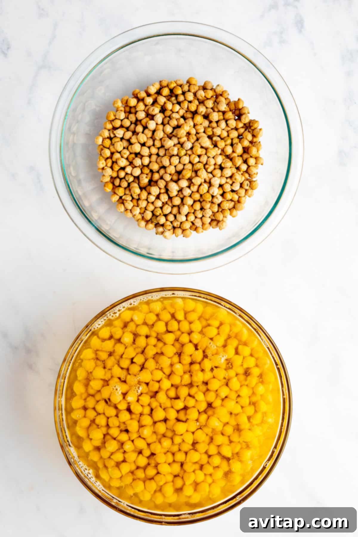 Two glass bowls on a marble surface: one with dried chickpeas and the other with soaked chickpeas in water.