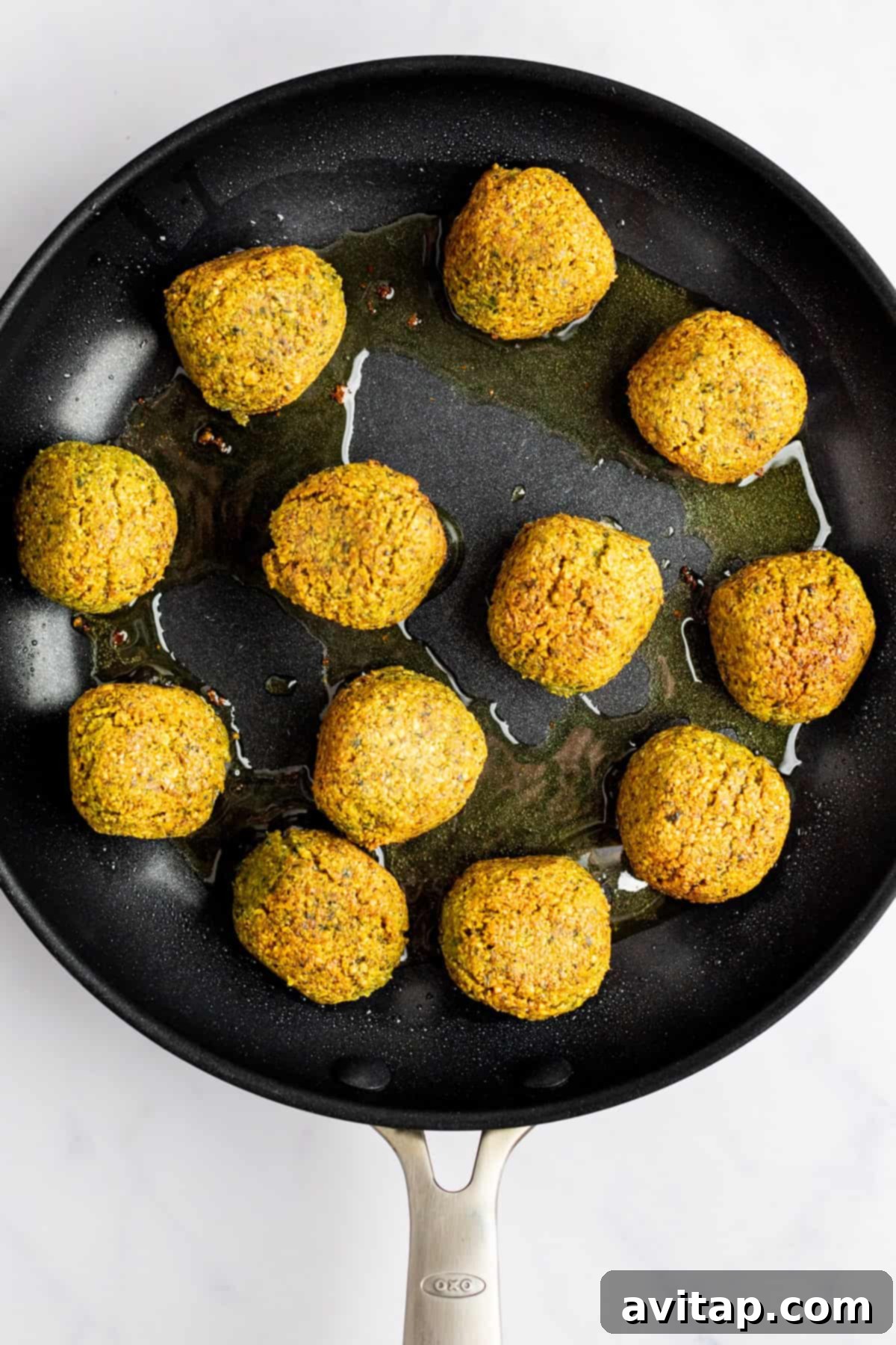 A skillet with twelve golden-brown falafel balls frying in oil on a light-colored surface.