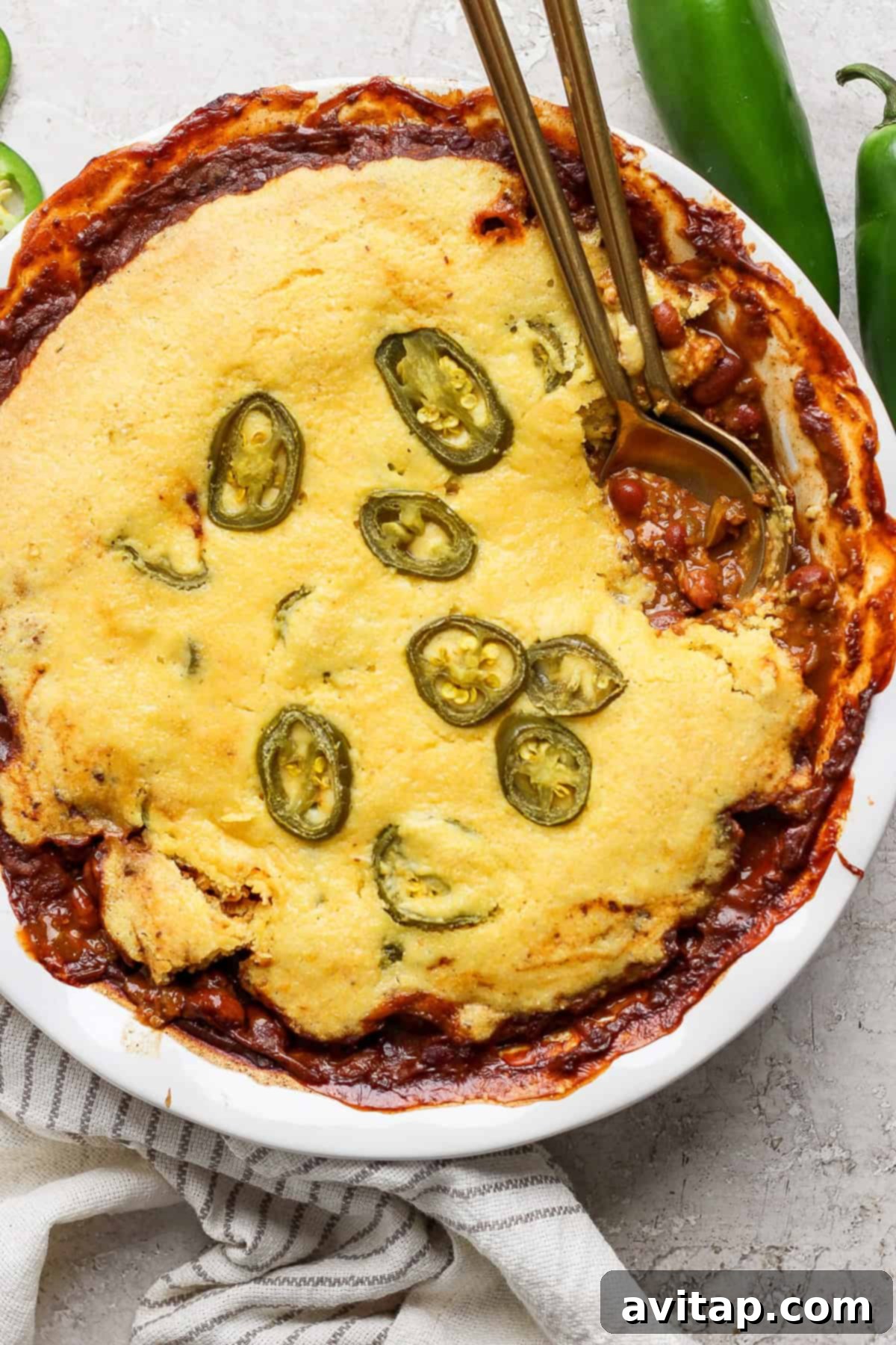 Hearty Chili Pot Pie 2 A golden-brown cornbread chili pie, generously topped with vibrant sliced jalapeños, presented in an elegant white pie dish. Two gold-colored spoons rest on the pie, all set on a light-colored surface, inviting diners to dig in.