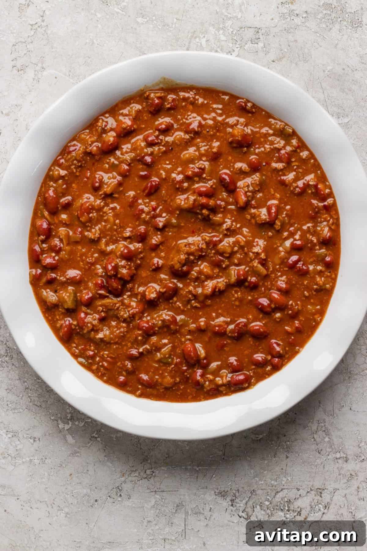 Hearty Chili Pot Pie 3 A sturdy white ceramic bowl brimming with a rich, dark red chili. The chili is packed with chunks of ground meat, kidney beans, and a thick, savory tomato-based sauce, all resting on a textured gray surface.