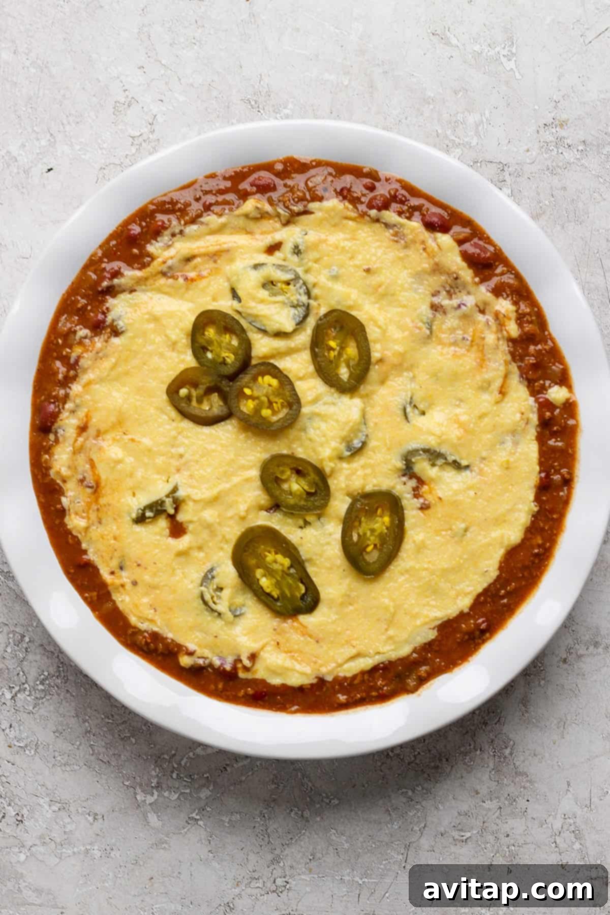 Hearty Chili Pot Pie 4 A close-up view of melted cheese bubbling over a rich red tomato-based sauce, generously garnished with bright green sliced jalapeños in a white dish, ready to be baked into a comforting chili pot pie.