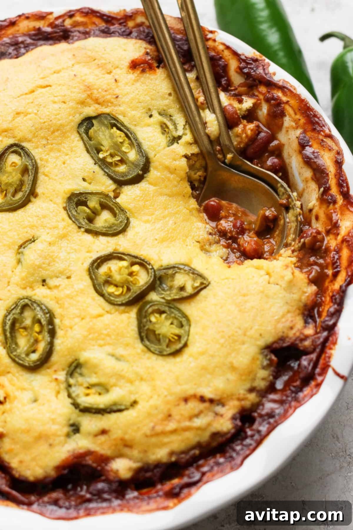 Hearty Chili Pot Pie 5 A perfectly baked chili pot pie in a round white ceramic dish, featuring a rich golden-brown cornbread topping generously sprinkled with vibrant green sliced jalapeños. The edges of the cornbread are delightfully crisp, and a peek of the savory chili filling is visible underneath. Two metal spoons are invitingly placed in the dish, ready for serving.