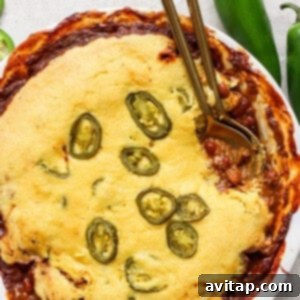 A baked chili pot pie in a round white pan, topped with cornbread and sliced jalapenos, with some edges browned and crispy.