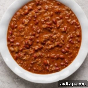 Hearty Chili Pot Pie 7 A white bowl filled with chili, containing ground meat, beans, and a thick tomato-based sauce, placed on a textured gray surface.
