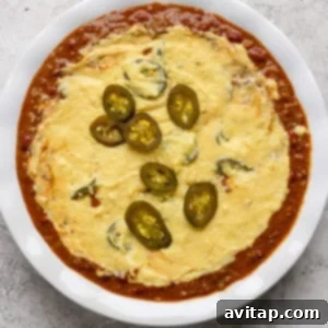 Hearty Chili Pot Pie 9 A white dish filled with melted cheese topped with sliced jalapeños, surrounded by a red tomato-based sauce.