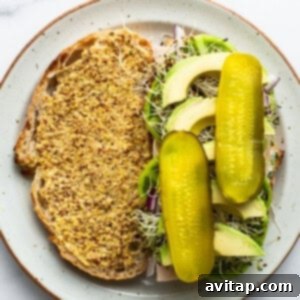 A plate of open-faced sandwich with mustard-covered bread, sprouts, sliced avocado, and two whole pickles on top.
