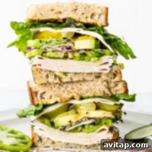 A tall sandwich with multiple layers of turkey, cheese, lettuce, cucumber, avocado, and sprouts on whole grain bread, served on a ceramic plate.