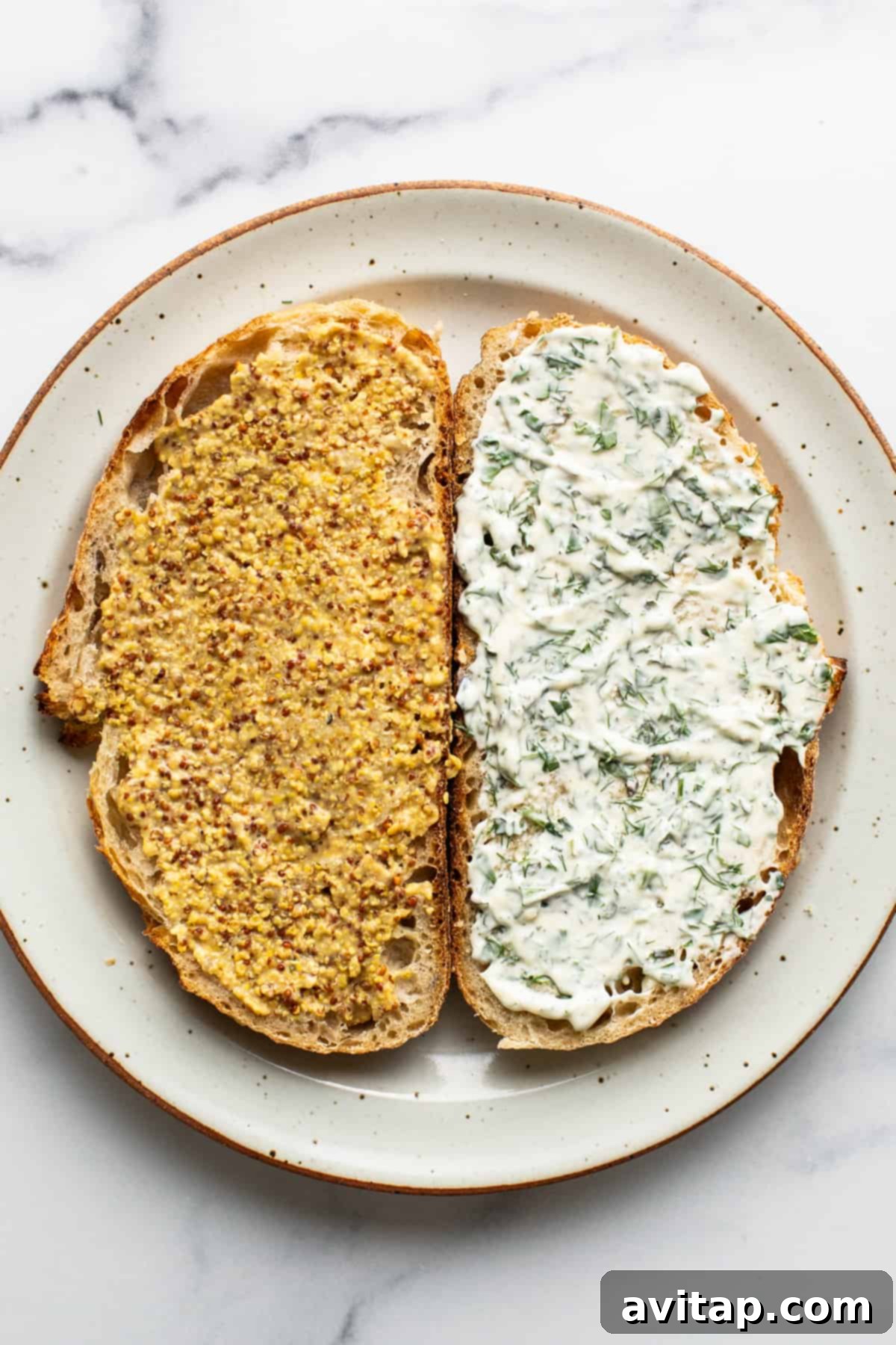 Two slices of bread on a plate, one topped with stone ground mustard, the other with creamy homemade herby mayo and chopped dill.