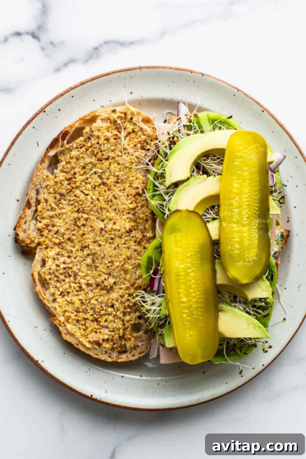A vibrant open-faced turkey sandwich showing layers of sprouts, creamy avocado slices, and whole deli pickles, all on mustard-covered bread.