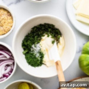 Overhead view of a kitchen counter with ingredients for a sandwich, including sliced cucumbers, cheese, pickles, red onions, and a bowl of mayonnaise mixed with herbs.