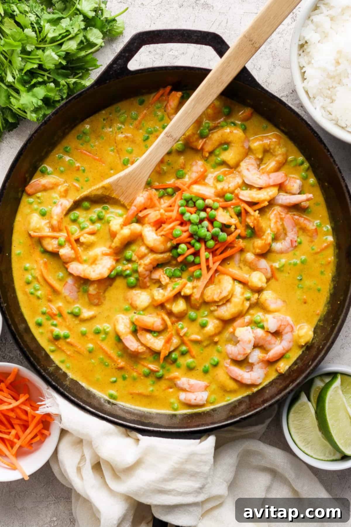 A skillet filled with vibrant shrimp and vegetable yellow curry, garnished with fresh green peas and served alongside fluffy jasmine rice, lime wedges, and aromatic fresh herbs, ready to be enjoyed.