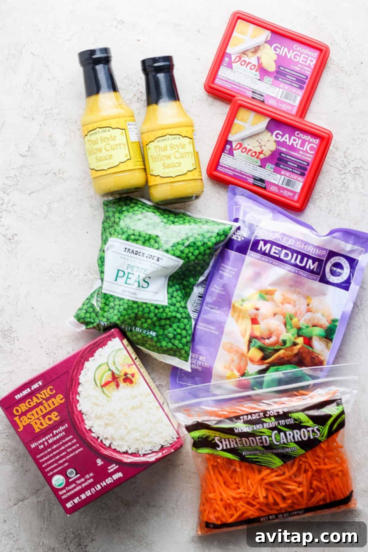 A vibrant flat lay showcasing all the essential Trader Joe's ingredients for a quick yellow curry, including bottles of Thai yellow curry sauce, a box of frozen ginger, packaged frozen garlic cubes, organic jasmine rice, a bag of petite frozen peas, and shredded matchstick carrots, all artfully arranged on a light textured surface.