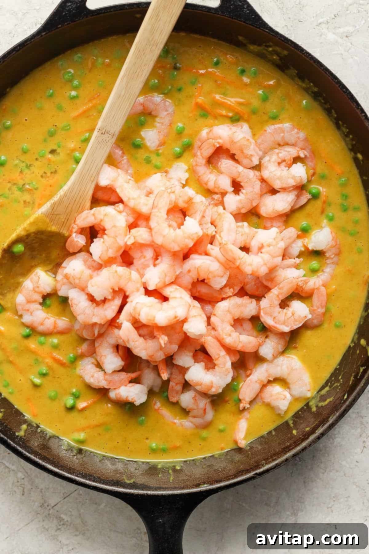 A close-up view of a deep skillet showcasing a luscious shrimp and pea curry, with a wooden spoon submerged in the creamy sauce, emphasizing its inviting texture and readiness to be served.