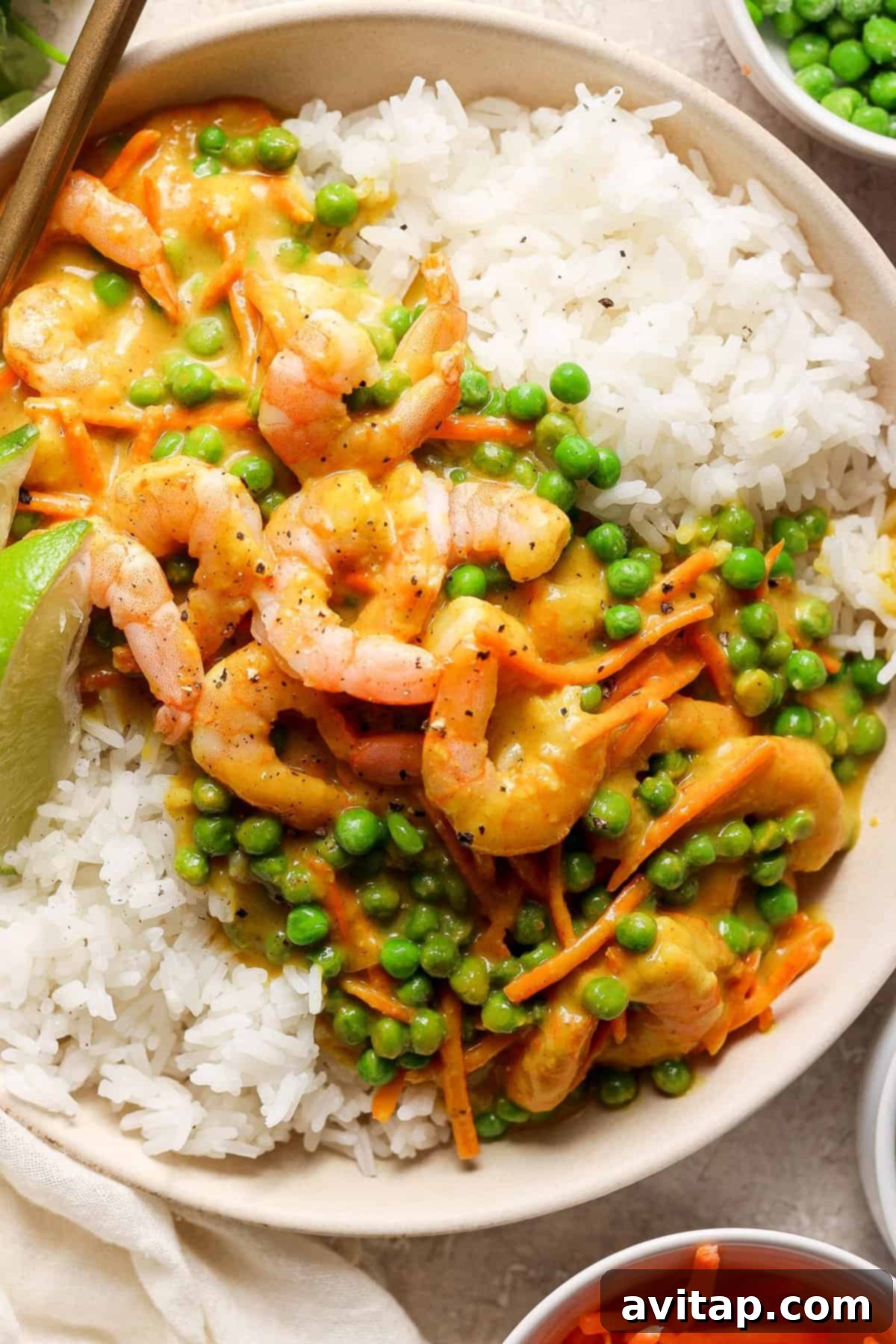 A beautifully plated bowl of fluffy white rice crowned with succulent shrimp and vibrant green peas, all bathed in a rich and creamy yellow curry sauce, elegantly garnished with a fresh lime wedge.