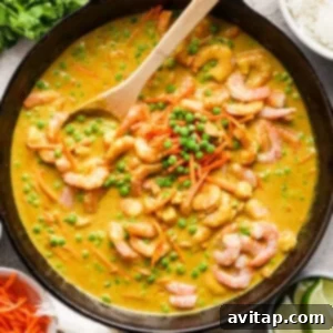 A wooden spoon is gently stirred into a deep skillet containing a vibrant shrimp curry with tender green peas and shredded carrots, accompanied by a side of fluffy white rice and fresh lime wedges, illustrating a delicious and complete meal.