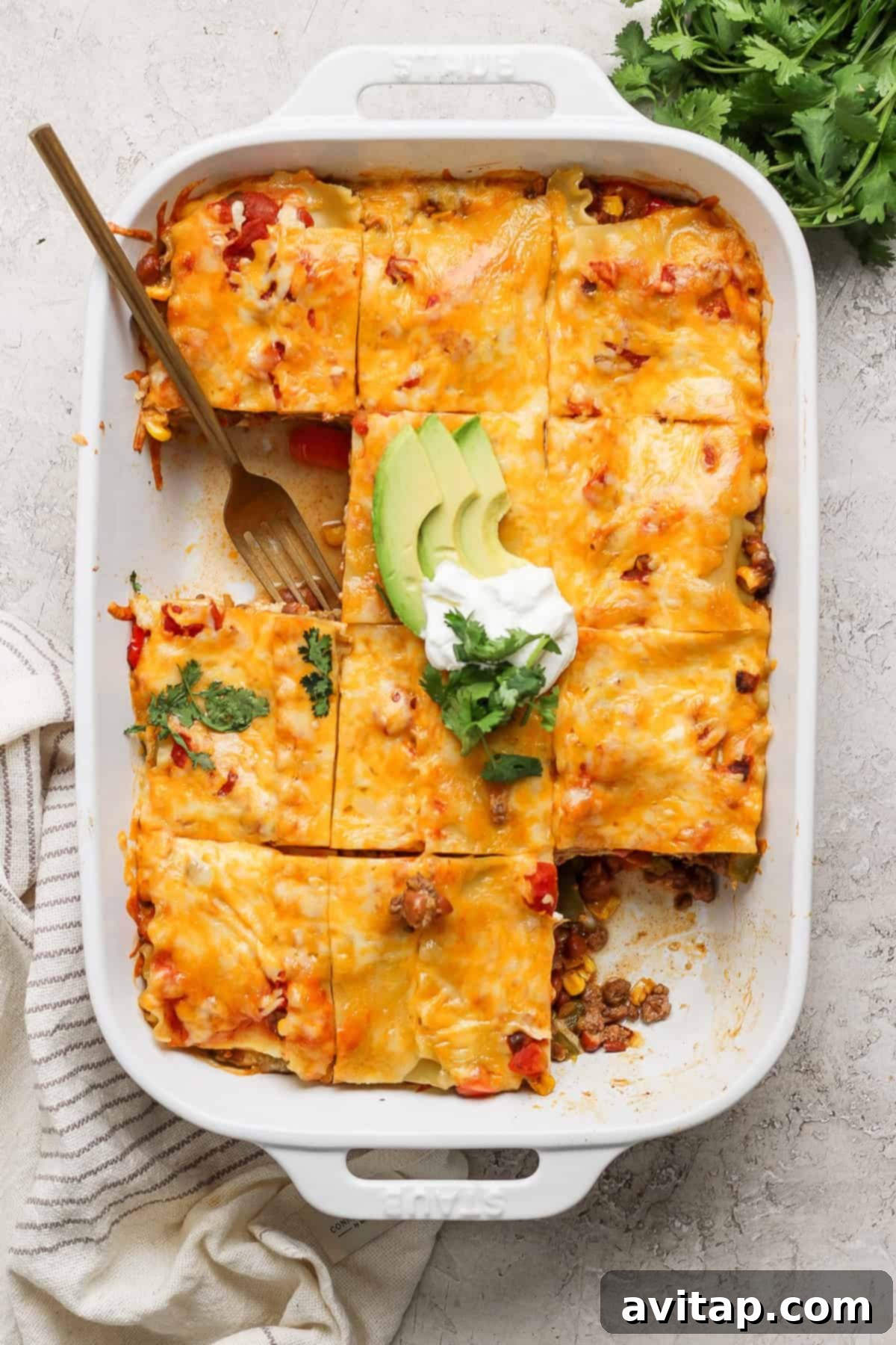 A baked casserole in a white rectangular dish is divided into squares, topped with melted cheese. Garnished with avocado slices, sour cream, and cilantro. A serving fork rests in one section, showcasing the layers of this delicious Whipped Cottage Cheese Taco Lasagna.