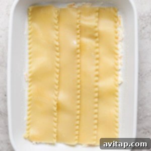 A white baking dish containing the first layer of four uncooked lasagna noodles laid flat on a bed of white sauce, preparing for the next delicious layer of taco lasagna.